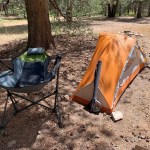 a small orange camping tent pitched on a dirt campsite with a foldable camping chair to its left the tent is set near a tree in a wooded area and there are rocks and dry ground around